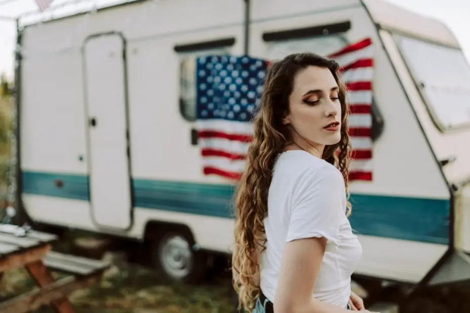 girl with long, curly brunette hair posing in front of an RV with an American flag hung on the side