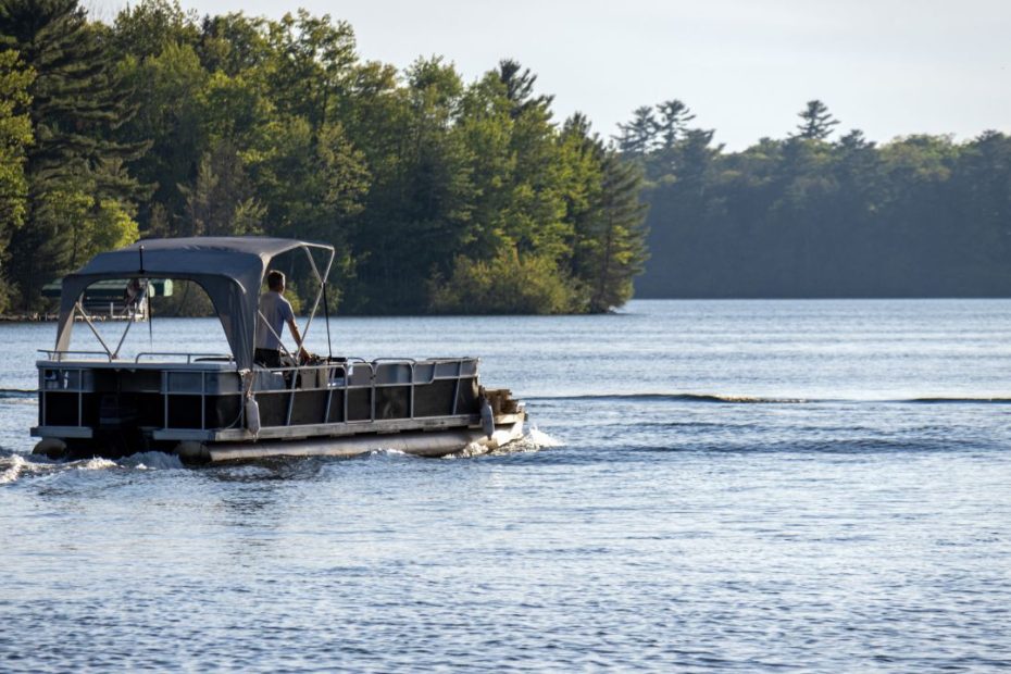 A man driving his pontoon boat in the middle of a lake with beautiful forest scenery in the background.