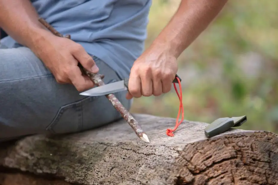 A man in jeans using a fixed-blade knife to whittle a stick he found while sitting on a log in the woods.