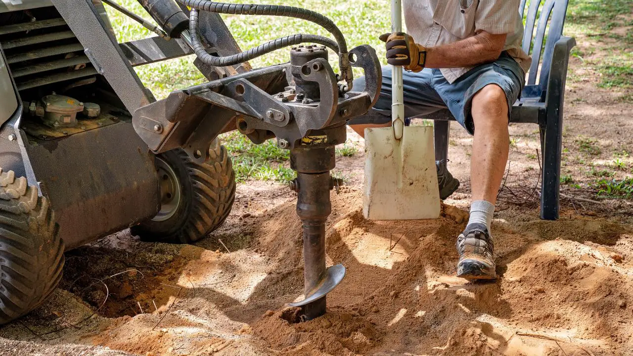 A man holds a shovel and sits in a chair as he uses a mechanical post hole digger for assistance in digging a hole.