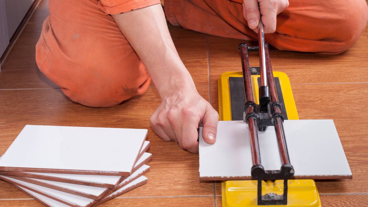 A close-up view shows a person using a yellow tool to cut a piece of white tile on a hardwood floor.