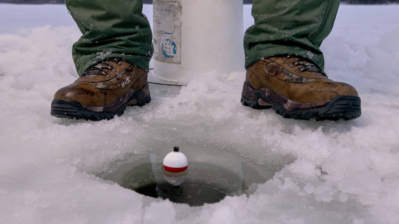 An angler sitting on a white bucket above a hole carved in ice with a fishing line and a bobber in the water.