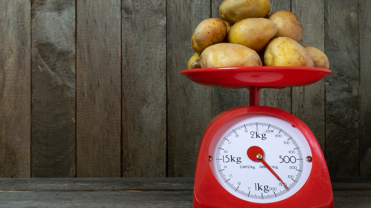 A stack of clean potatoes on a bright red mechanical scale on a dark wood table with a dark wood vertically paneled wall.