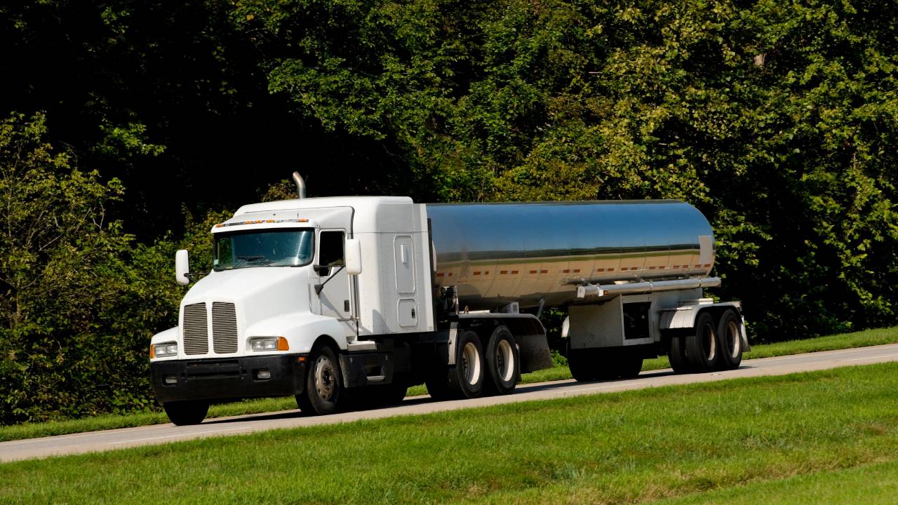 A stainless steel tanker trailer with a white cab drives down a road next to grass with several trees in the background.