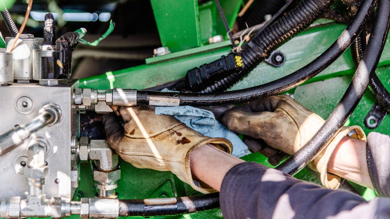 Close-up of a person reaching into the undercarriage of an industrial harvester, grabbing onto hydraulic hoses.