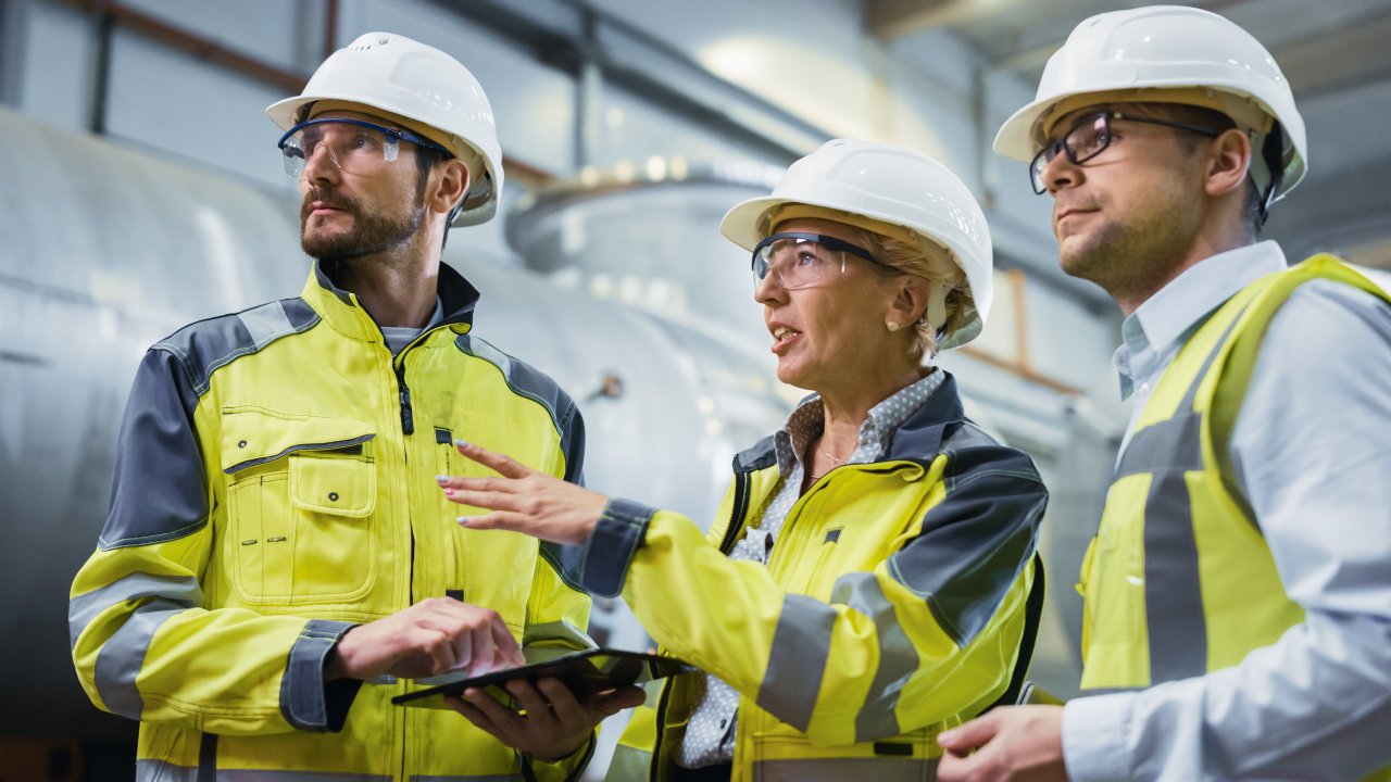 Three oil and gas workers wearing white hard hats, yellow vests, and glasses stand together on-site