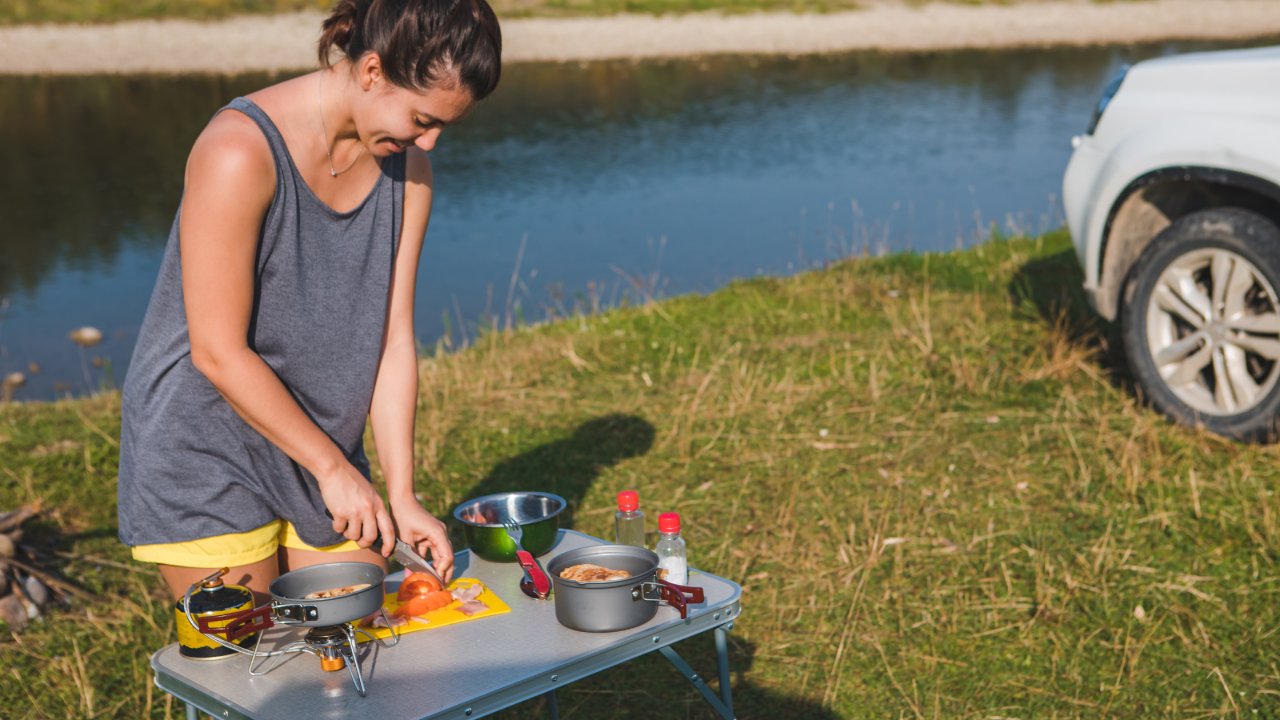 A woman wearing a gray tank top and yellow shorts is preparing a meal outside. She is standing next to a body of water.