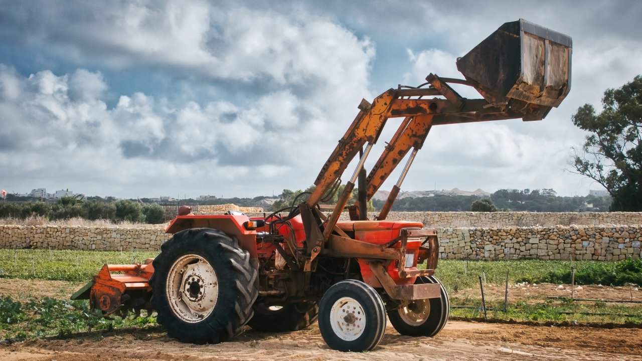 A red tractor with a raised front loader parked on a cleared section of land near a stone wall. There are trees in the distance.