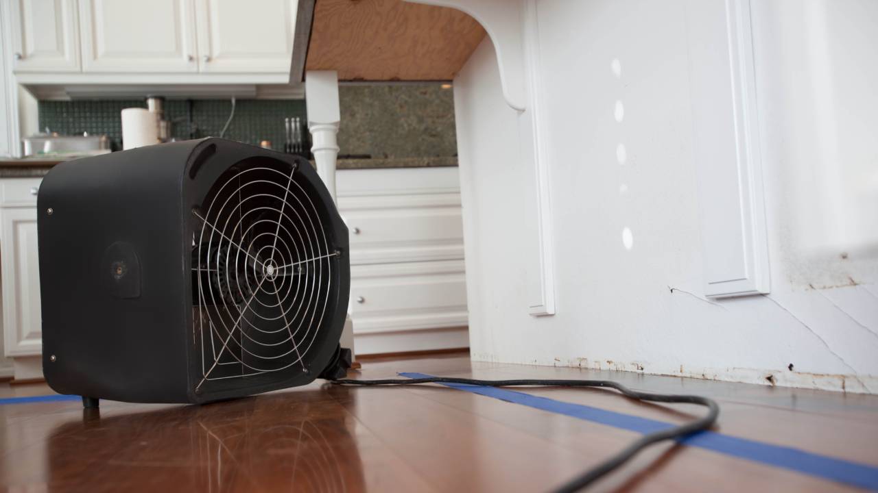 White kitchen cabinets with removed baseboards are being dried by a square black industrial fan to repair water damage.