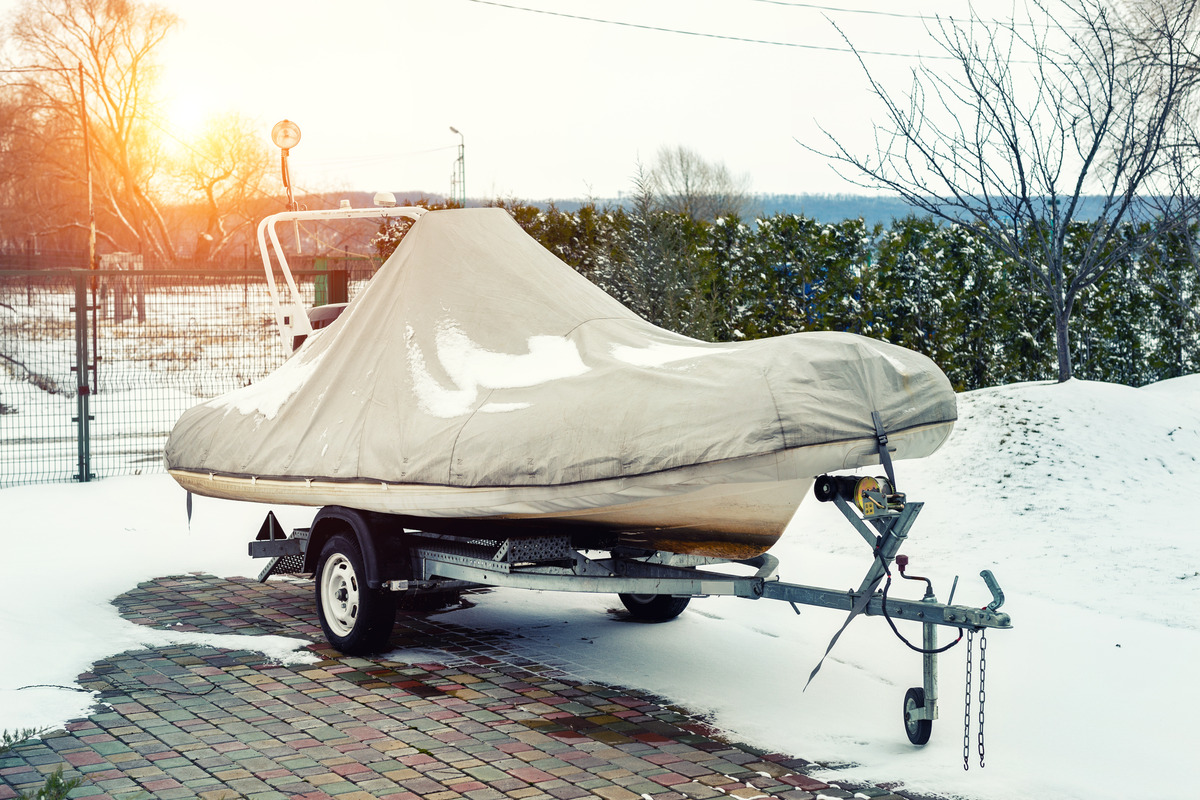 A winterized small motorboat covered and stored on a trailer in a brick-paved driveway, with snow covering the ground.