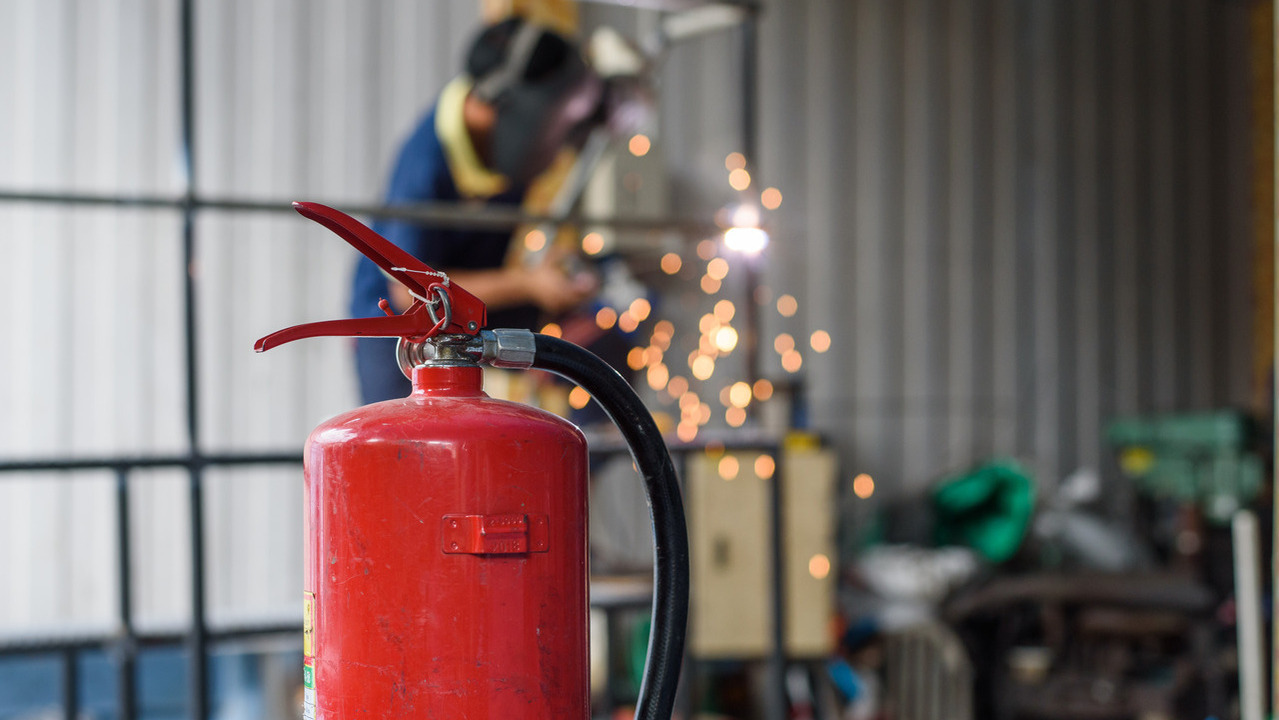 A fire extinguisher is ready for use as a person works on a welding project in the background, emitting hot sparks.