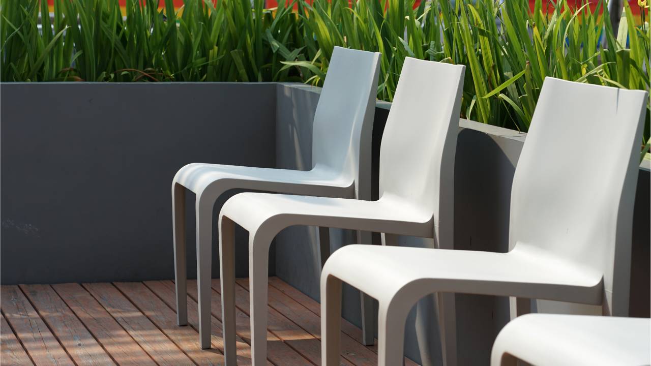 In an outdoor area with soft sunlight, four grey chairs are lined up by a grey planter box with green vegetation.