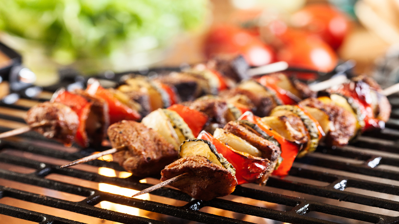 Close-up of three skewers with meat and different vegetables placed on a metal grate with a flame underneath.
