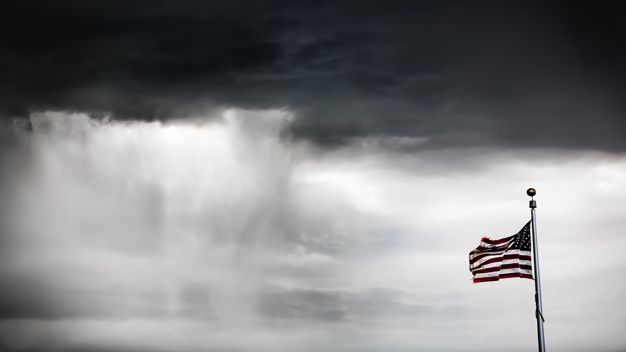 A large American flag hanging on a flagpole blows in the wind as dark storm clouds fill the background.