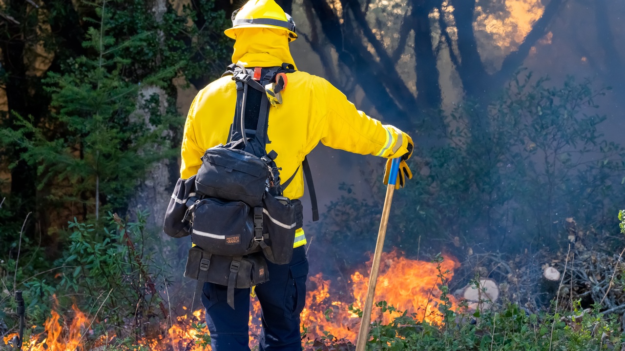 A wildland firefighter wearing a protective red hard hat and yellow suit stands while watching a wildfire burn.