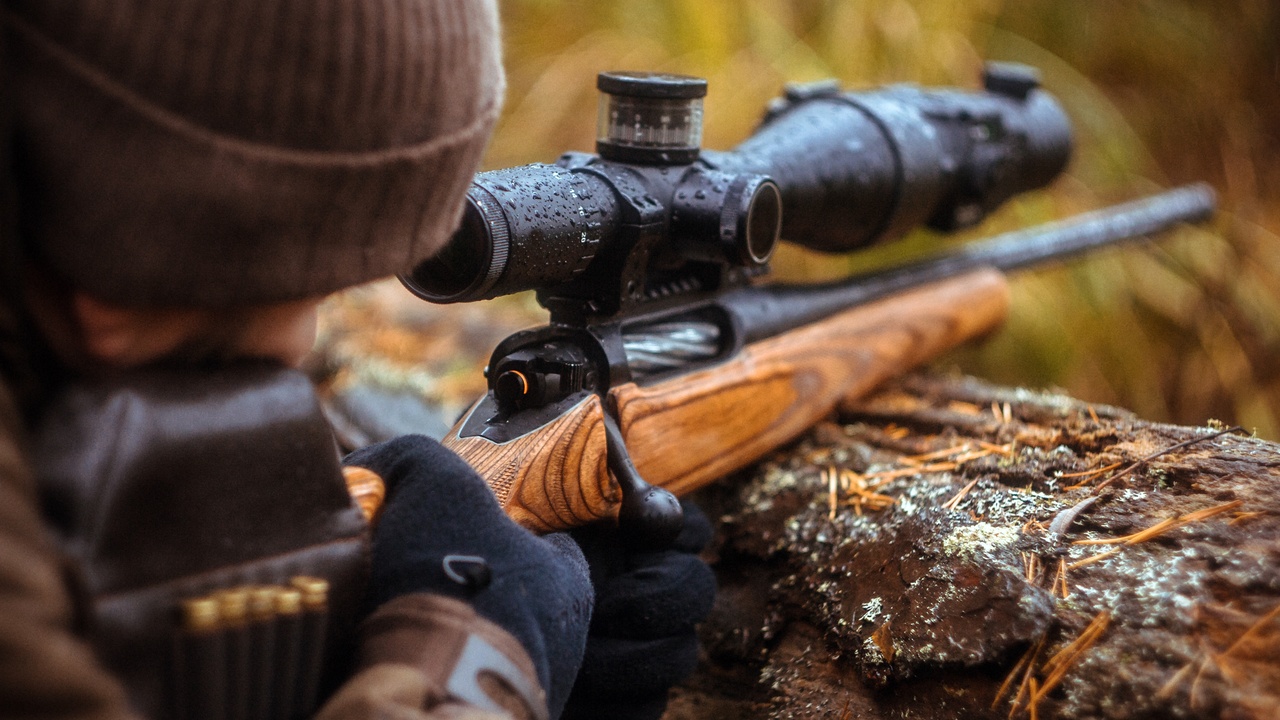 A person wearing a brown jacket and hat uses a log to steady a rifle. They are looking through the gun scope.