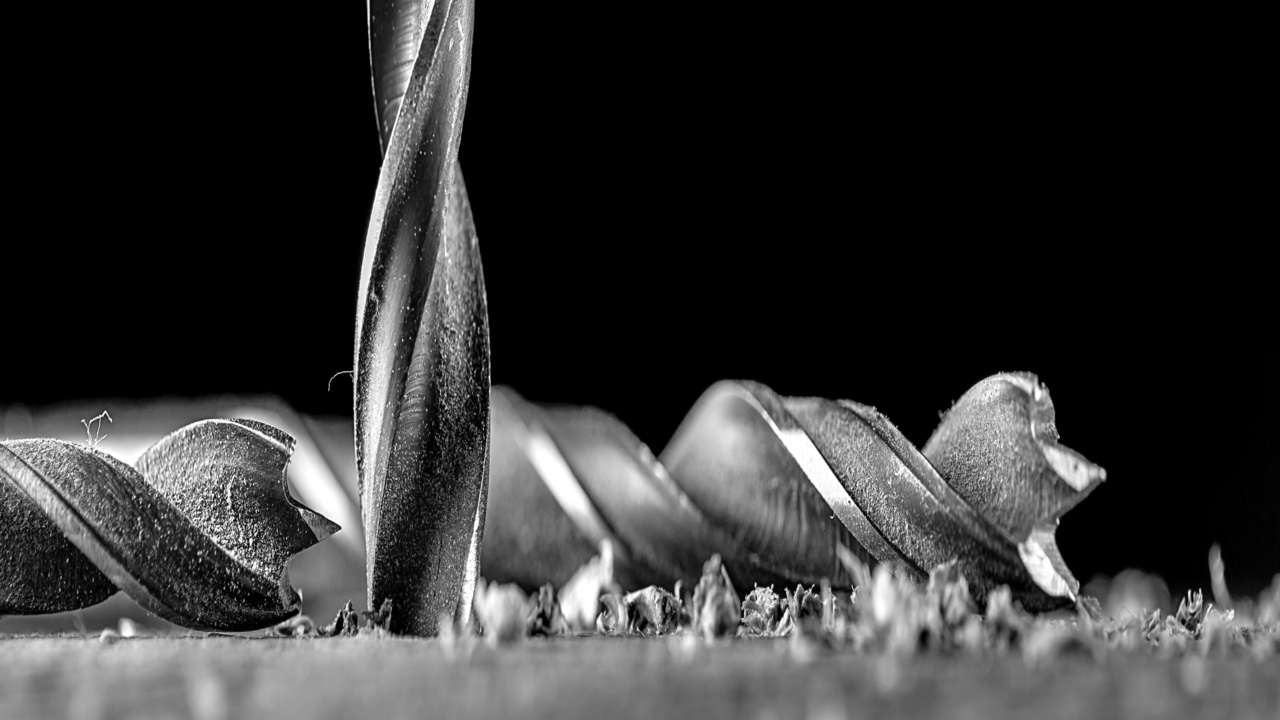 An extreme close-up view shows a silver drill bit drilling into a piece of metal. Another bit lies beside the other bit.