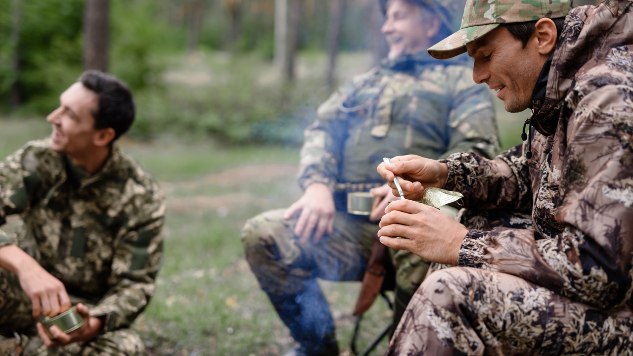 Three men wearing camouflage hunting gear sit around a bonfire in a forest and eat food out of tin cans.