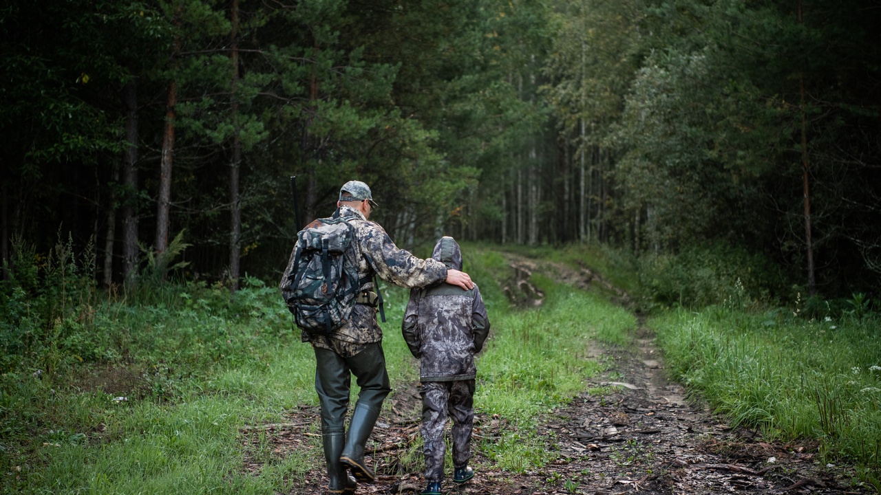 A grown man and a young boy are both wearing full camouflage clothing as they walk down a trail into a forest.