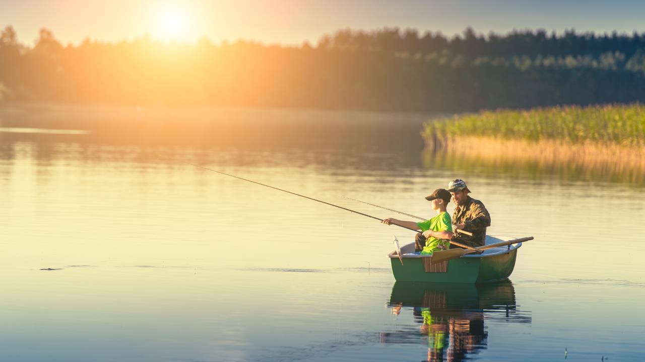 A father and a son wearing baseball hats sit in a green boat holding fishing poles. The sun is setting behind trees.