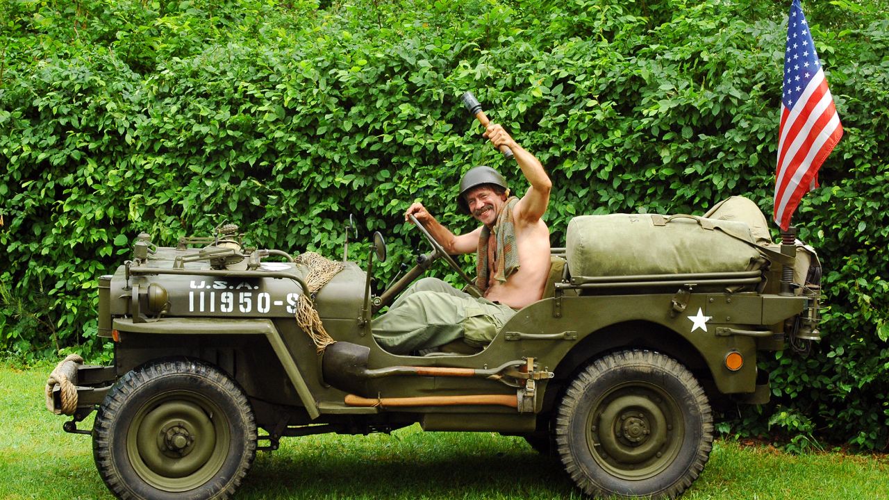A man wearing a helmet sits in a vintage World War II era Willys military Jeep that is dark green in color.
