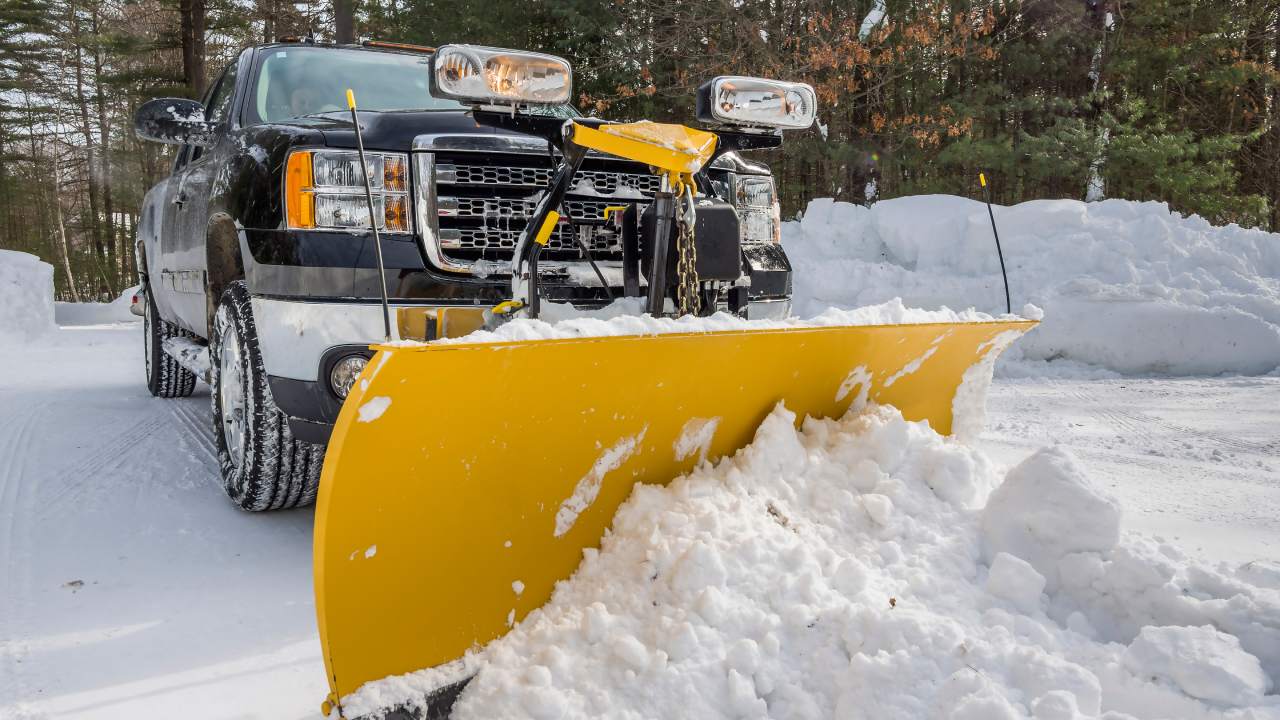 A dark-blue colored pickup truck has a yellow snowplow attached to the front. It sits in a snowy driveway.