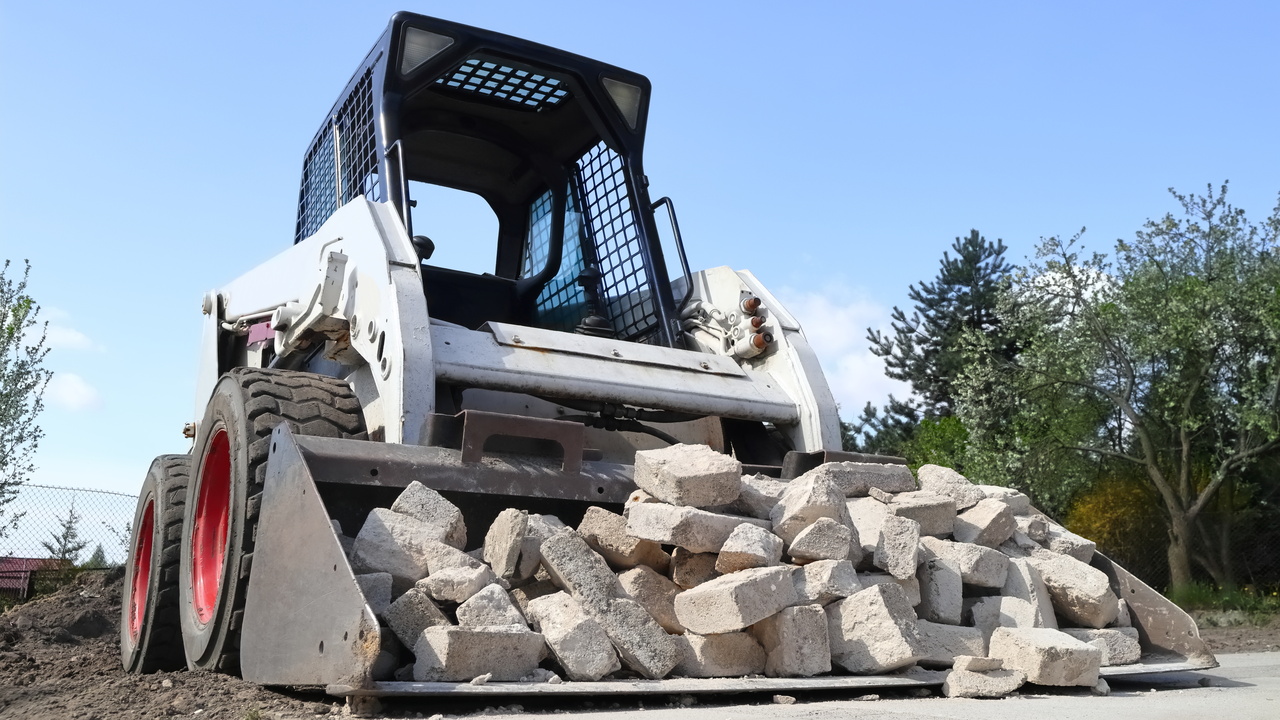 A white skid steer has a black bucket attachment on the front that is filled with gray bricks. The skid steer sits on dirt.