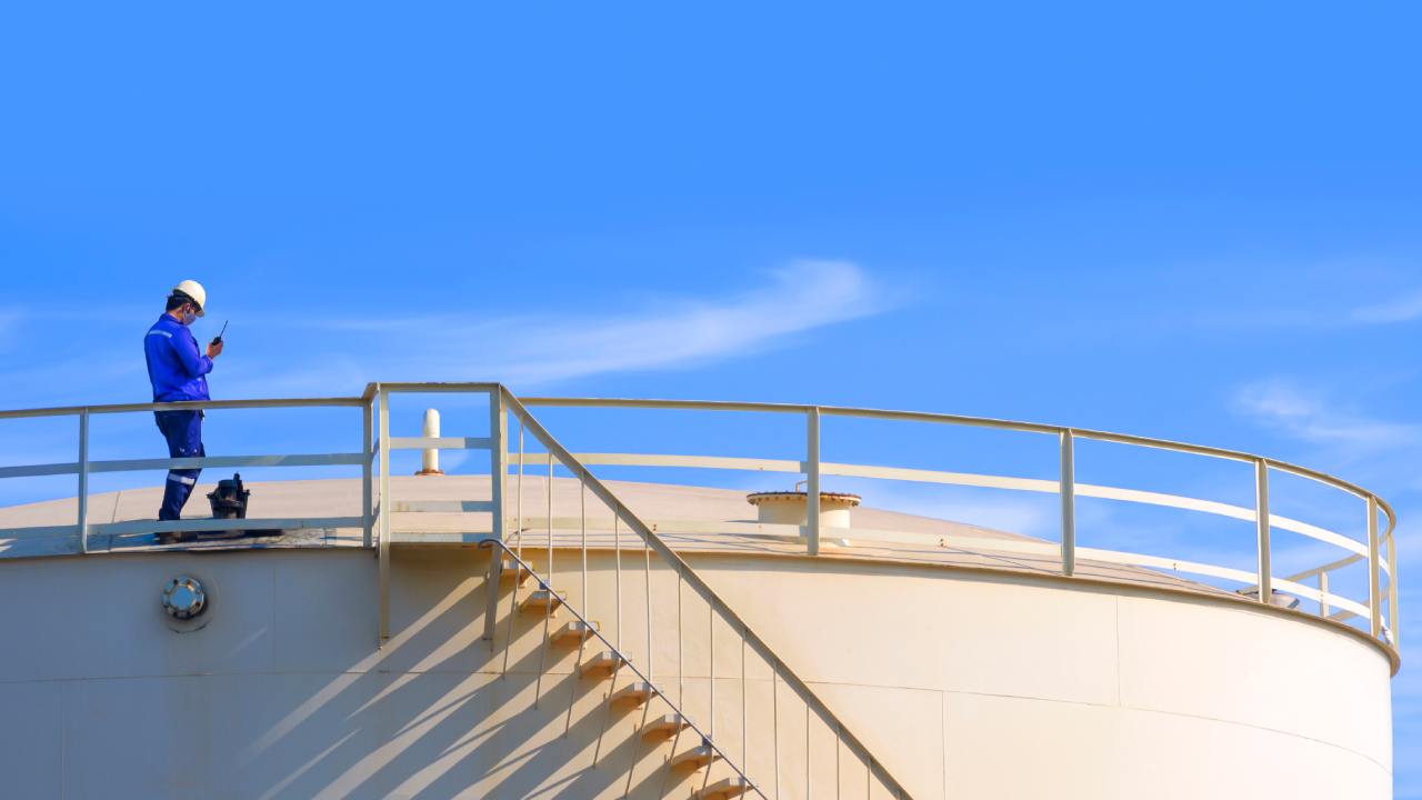 A person wearing a bright, blue jacket and a white hard hat holds a walkie-talkie while standing on a white industrial tank.