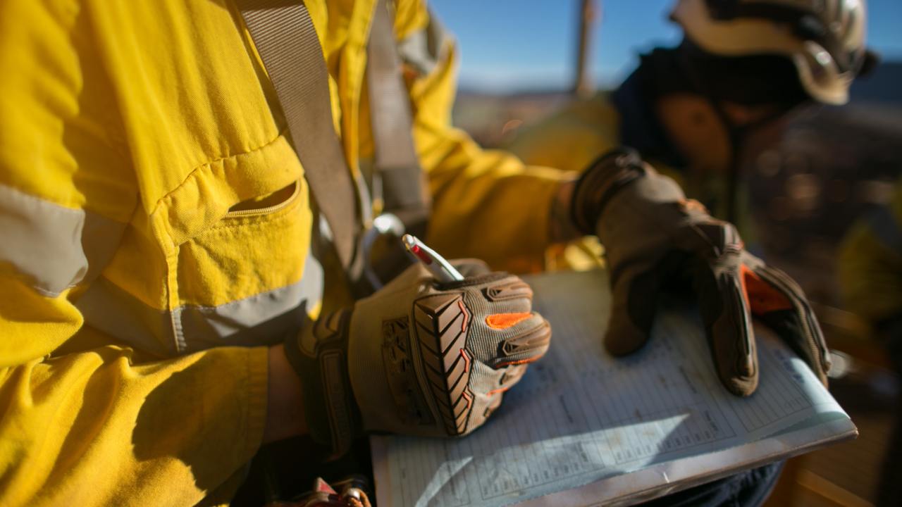 A person at work wears a yellow safety jacket and a pair of safety gloves. They're writing on a clipboard.