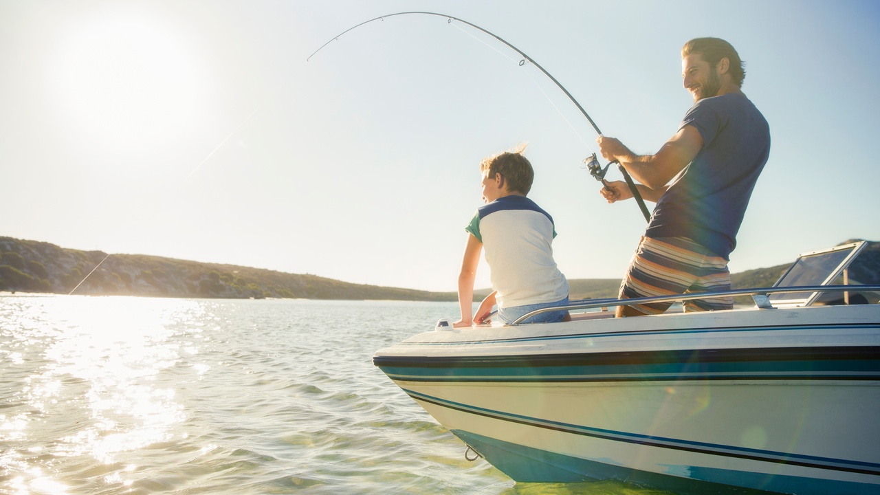 On a sunny day, a smiling father and his young son fish from the bow of a blue, white, and black boat.