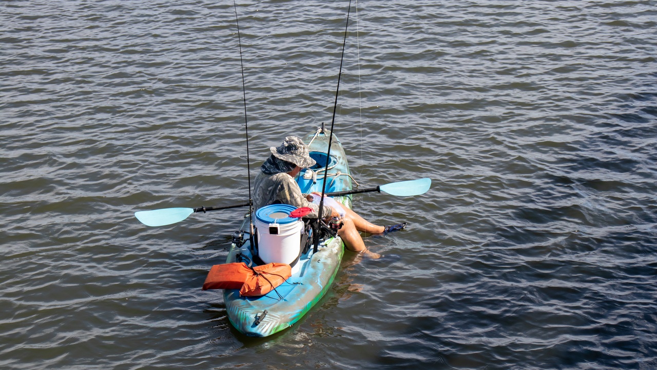 Someone sits in their kayak with their feet hanging in the water. Fishing gear and a life jacket are next to them.