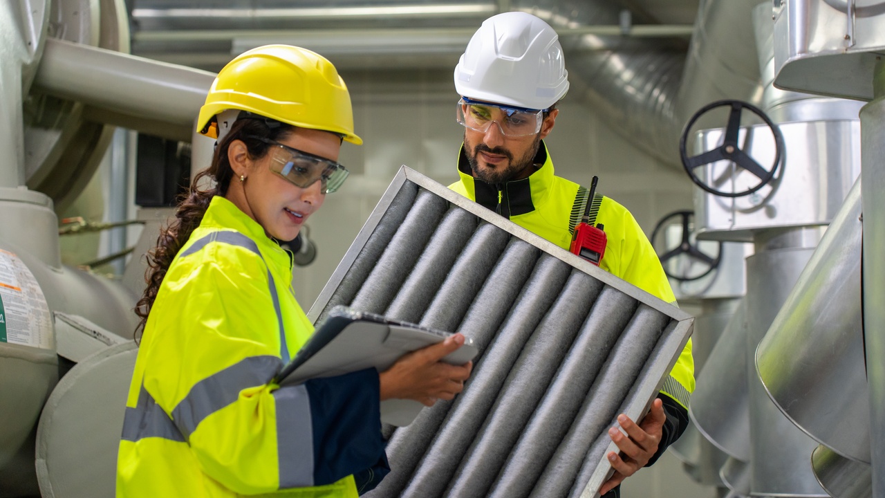 A man and a woman worker wearing safety gear in an industrial setting. The woman holds a clipboard and the man holds an air filter.