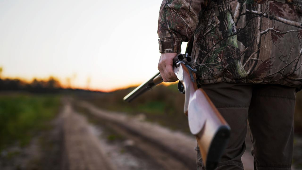 A hunter stands on a dirt trail outdoors, holding a rifle at his side with the loading side open and visible.