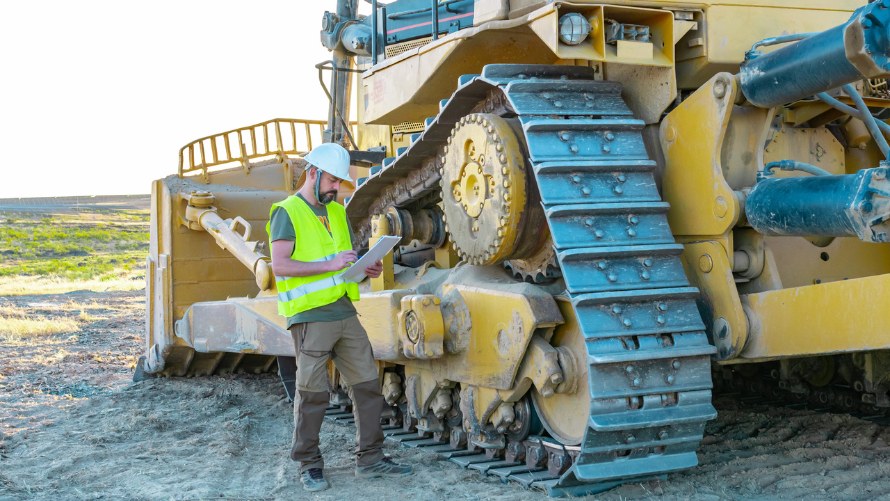 A man wearing a yellow reflective vest and a white hard hat stands at the base of a bulldozer. He takes notes on a clipboard.
