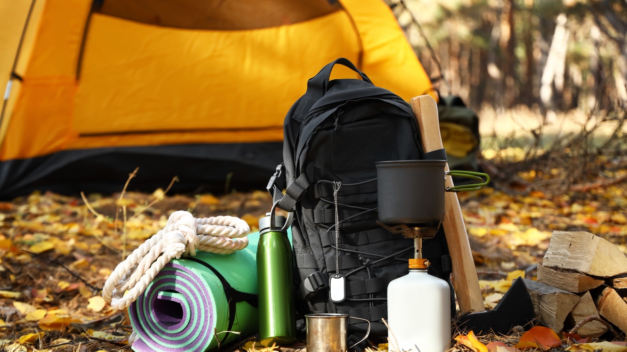 A set of camping gear, with metal mugs, a white rope, and a portable stove sit on the ground next to a tent.