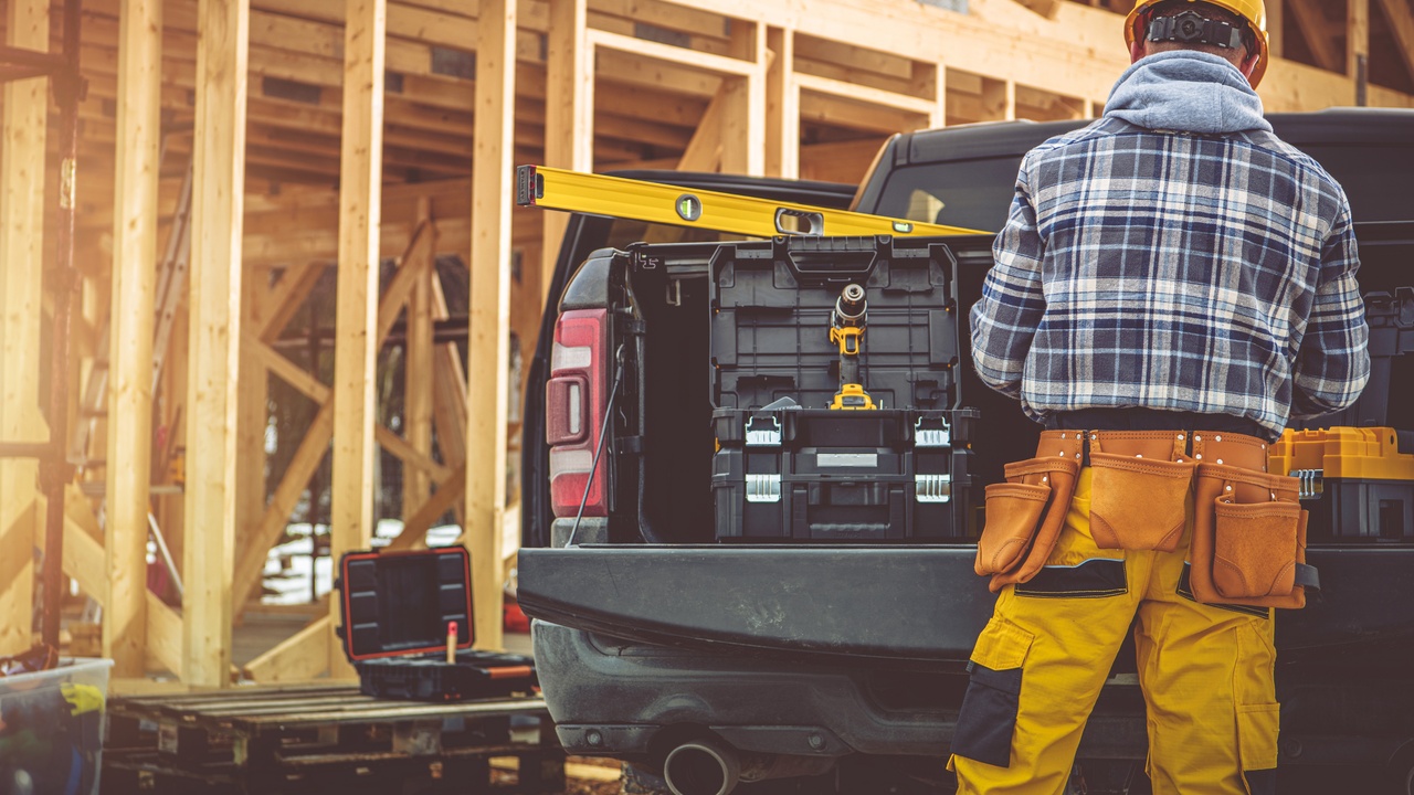 A man wearing a flannel shirt, a helmet, and a tool belt, standing behind a pickup truck at a construction site.