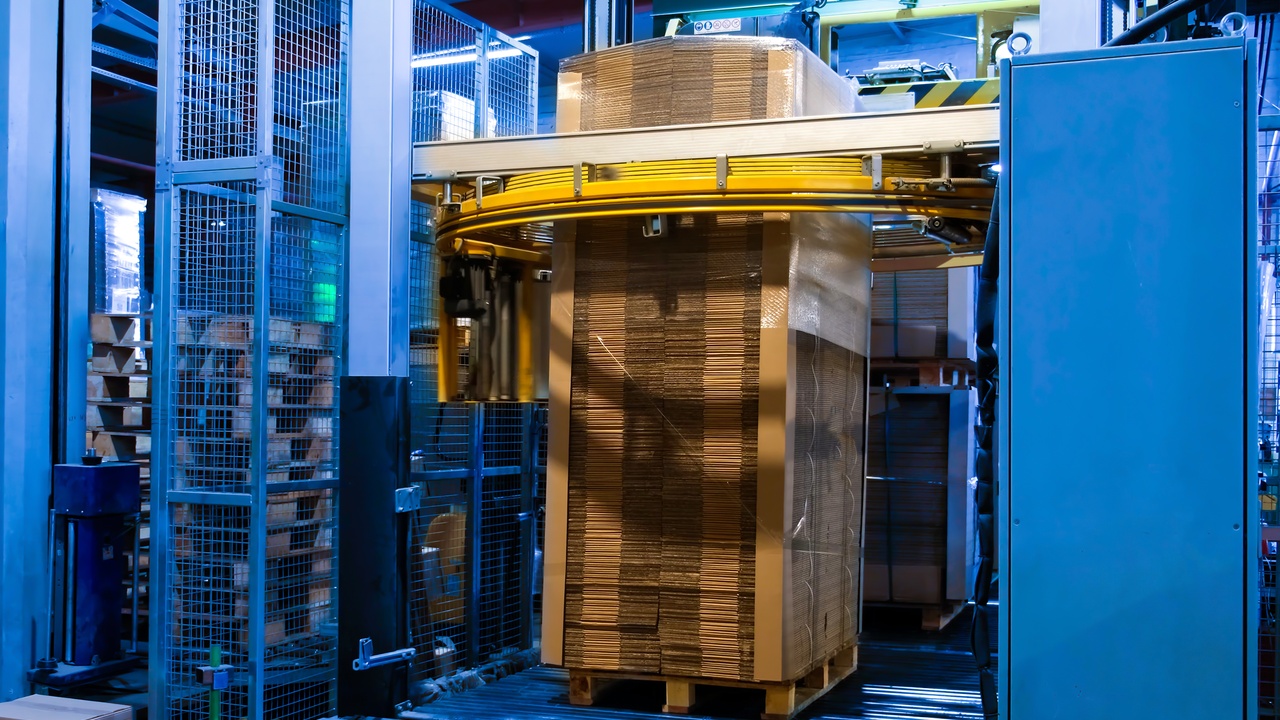 An automated pallet wrapping machine securing a tall stack of cardboard boxes on a pallet in a warehouse.