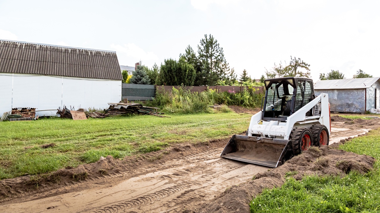 A white skid steer with a bucket attachment parked on a dirt path near a few buildings. There is grass on each side of the path.