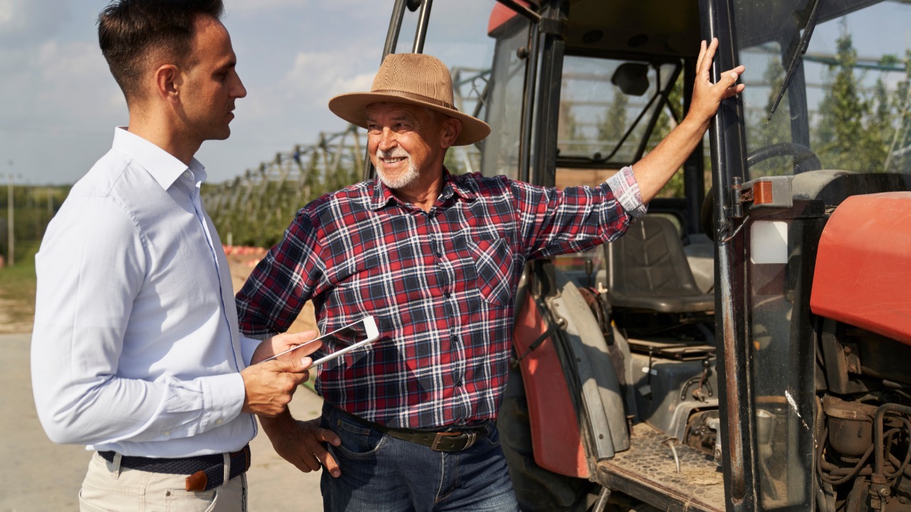 A senior farmer resting his hand on the frame of a red tractor as he chats with a man holding a tablet.