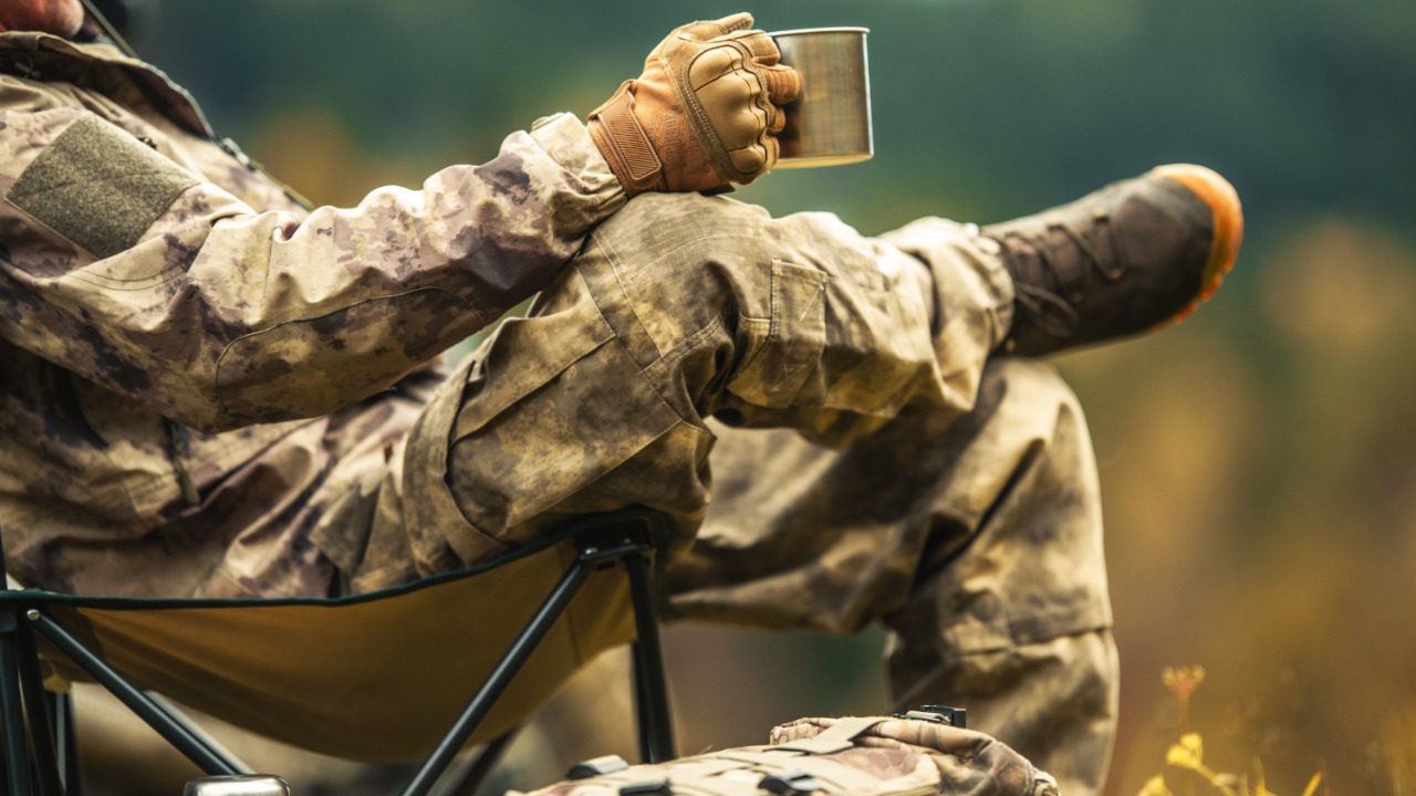 A solo hunter sitting on a camping chair and looking out to the wilderness while drinking from a metal cup.